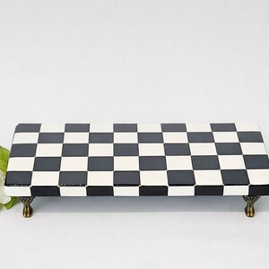 May include: A rectangular riser featuring a black and white checkered pattern on its top surface. The platform is supported by four small, ornate, metallic bronze-toned feet. A cluster of white flowers with green leaves is positioned to the left side.