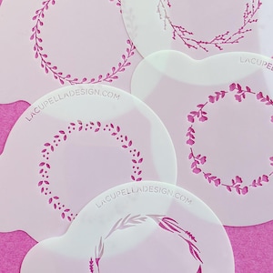 May include: Several white stencils featuring floral wreath patterns in pink hues. The stencils are displayed on a pink background. Some stencils have the website address LACUPELLADESIGN.COM printed on them.