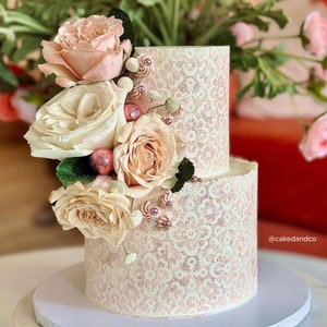 May include: Two-tiered white wedding cake with a delicate lace design and a blush pink hue. The cake is adorned with a cluster of blush and ivory roses, greenery, and gold accents.