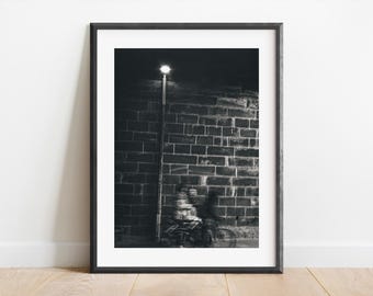 May include: Black and white photograph of a person riding a bicycle under a streetlight next to a brick wall.