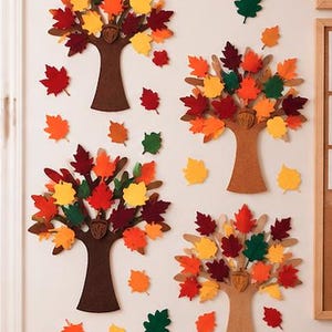 May include: Four decorative wall cutouts shaped like trees with brown trunks and colorful felt leaves in shades of red, orange, yellow, and green. Each tree features a small brown acorn icon at the base of the canopy, with additional loose leaf shapes scattered around the trees on the wall.