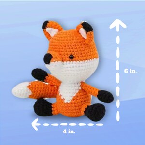 Op de afbeelding: Een gehaakt oranje en wit pluche vosje met zwarte ogen en een zwarte neus. Het speelgoed zit op een blauwe achtergrond en is 15,24 cm hoog en 10,16 cm breed.