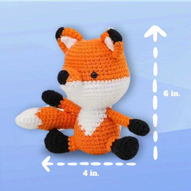 Op de afbeelding: Een gehaakt oranje en wit pluche vosje met zwarte ogen en een zwarte neus. Het speelgoed zit op een blauwe achtergrond en is 15,24 cm hoog en 10,16 cm breed.