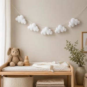 May include: A nursery scene with a wooden changing table, a woven basket, and a stack of blankets. A plush bunny and wooden stacking rings sit on the table. A garland of white cloud decorations hangs above. A vase with greenery and a framed picture complete the scene.