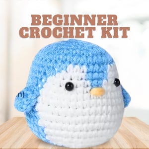 May include: Beginner crochet kit featuring a blue and white penguin amigurumi. The penguin has black eyes and an orange beak.