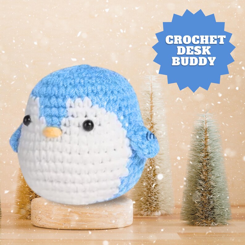 May include: A crocheted penguin desk buddy in blue and white. The penguin is sitting on a wooden base. The image has a snowy background with small pine trees.
