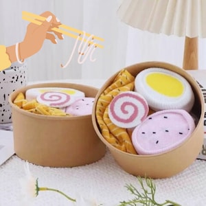 May include: Two brown paper bowls filled with socks designed to look like ramen. One bowl has a slice of egg, a pink swirl, and a pink sock with black dots. The other bowl has similar items. A hand with chopsticks is holding up noodles.