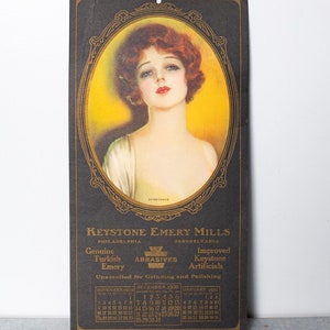 May include: A vintage calendar card for Keystone Emery Mills, featuring a woman with red hair in a gold frame. The calendar shows the months of November, December, and January, with the years 1930 and 1931. The text on the card reads "Keystone Emery Mills, Philadelphia, Pennsylvania, Genuine Turkish Abrasives, Improved Keystone Artificials, Unexcelled for Grinding and Polishing."