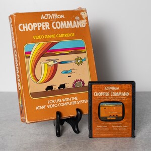 May include: A vintage Atari video game cartridge for the game "Chopper Command" by Activision. The box art features a helicopter flying over a desert landscape with a rainbow in the background. The cartridge is black with a label that says "Chopper Command" and has a picture of a helicopter flying over a desert landscape.