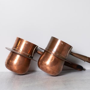 May include: Two antique copper coffee pots with wooden handles. The pots have a round body and a flat base. The handles are attached to the side of the pots. The pots are marked with the number "112".