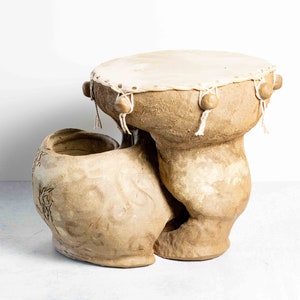 May include: A beige, handcrafted drum with a light-colored drumhead. The drum has a unique shape with a rounded base and a curved, decorative side. Wooden beads and white cords adorn the top.