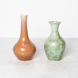 May include: Two ceramic vases. One is a tall, slender vase with a brown and peach gradient. The other is a shorter, round vase with a green, crystalline pattern. Both vases have a glossy finish and are set against a neutral background.