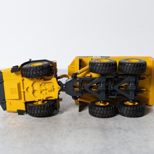May include: A yellow toy dump truck with black wheels. The truck is tipped over on its side, showing the underside of the chassis.