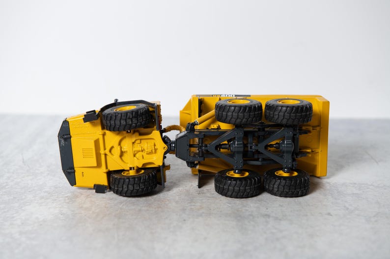 May include: A yellow toy dump truck with black wheels. The truck is tipped over on its side, showing the underside of the chassis.