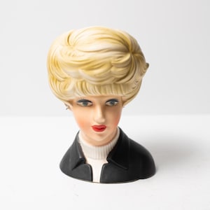 May include: A ceramic head planter featuring a stylized face with blue eyes, red lips, and blonde hair styled in a voluminous bob. The figure wears a black collared jacket over a white ribbed turtleneck sweater, creating a detailed bust sculpture.