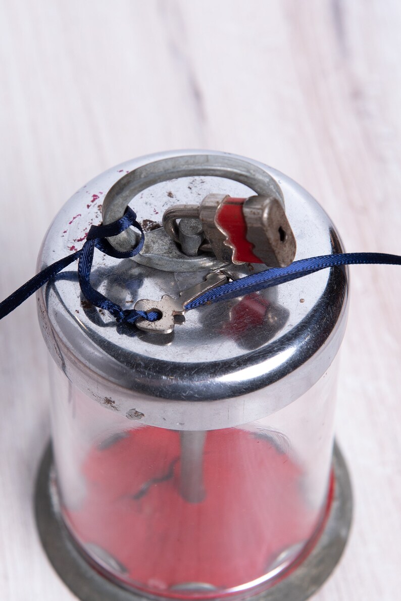 May include: A silver and glass piggy bank with a red interior. The lid is secured with a small padlock and a blue ribbon. A key is attached to the padlock.
