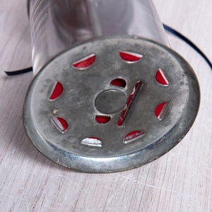 May include: A vintage metal and glass spice grinder with a red and silver design. The grinder has a circular base with a hole in the center and a glass jar on top.