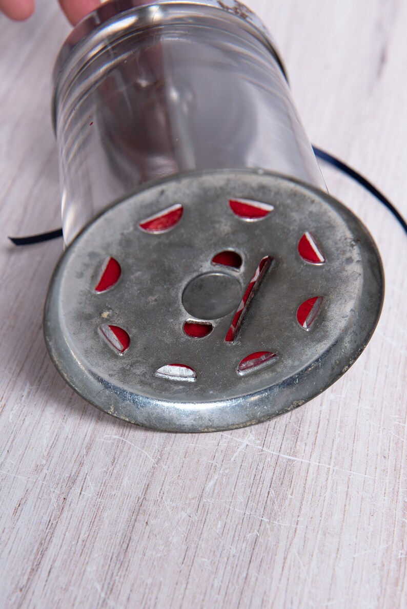 May include: A vintage metal and glass spice grinder with a red and silver design. The grinder has a circular base with a hole in the center and a glass jar on top.