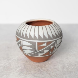 May include: A round, hand-painted ceramic pot with a wide opening. The pot features a geometric design in white, grey, and terracotta colours. The interior is terracotta. The pot is a decorative item.