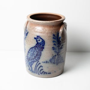 May include: A cylindrical stoneware crock featuring a tan, salt-glazed finish with two side handles. The exterior is decorated with cobalt blue hand-painted motifs of a bird and stylized foliage, along with an impressed oval maker's mark.