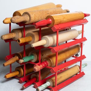 May include: A red metal rack holding several wooden rolling pins. The rolling pins are made of natural wood with red, green, and yellow handles. The rack is a grid, designed to store the rolling pins horizontally. The rack is painted red.