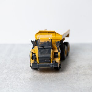 May include: A yellow toy dump truck with a black cab and black wheels. The truck is on a gray surface.