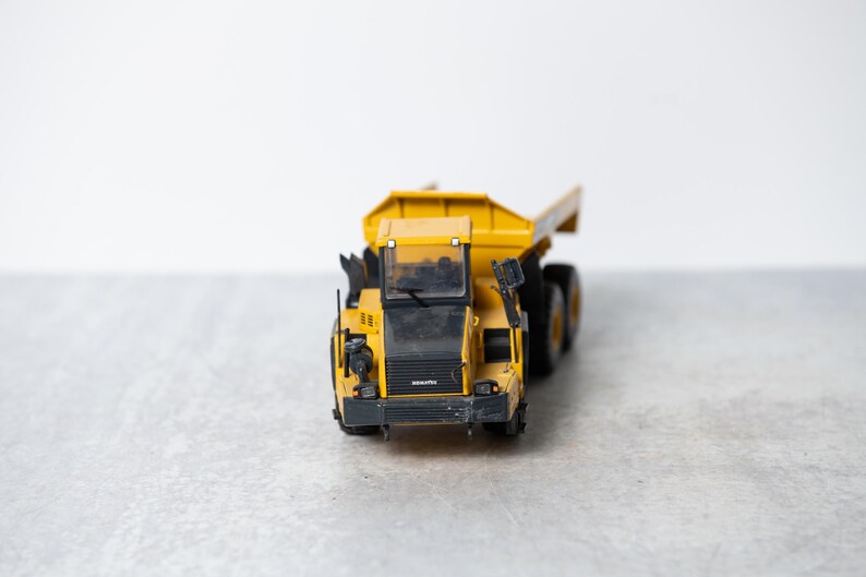 May include: A yellow toy dump truck with a black cab and black wheels. The truck is on a gray surface.