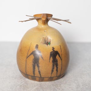 May include: A handcrafted ceramic vase with a rounded body and a narrow neck. The vase is tan with black silhouettes of figures and a decorative twig arrangement. A small green bead is attached to the vase.