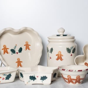 May include: A collection of off-white ceramic kitchenware with gingerbread man and holly designs. Includes a heart-shaped pie dish, a jar with a lid, a heart-shaped bowl, and various rectangular and star-shaped dishes.