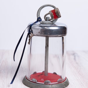 May include: A vintage glass jar with a metal lid and a red padlock. The jar has a metal rod running through the center and a red base. A blue ribbon is tied around the lid with two keys attached.