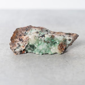 May include: A rough, brown rock with a cluster of small, green and white crystals. The crystals are clustered together in a dense formation.
