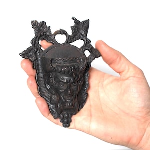 May include: A dark, cast iron wall hanging featuring a detailed face with a laurel wreath and grape vine accents. The decorative piece is held in a hand against a white background. The face has a stern expression and is surrounded by ornate leaves.