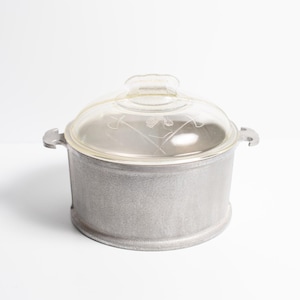 May include: A small, round, silver-colored cooking pot with a clear glass lid. The lid has a decorative etched design. The pot has two side handles and is sitting on a white surface.