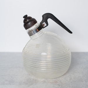 May include: Vintage clear glass seltzer bottle with a black handle and a dark brown top. The round bottle has horizontal ridges and a metal band around the neck. The handle is angled, and the top has a small nozzle.