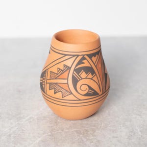 May include: A terracotta-colored ceramic vase with a rounded base and a wide opening. The vase is adorned with a black and orange geometric pattern featuring triangles, lines, and a spiral design. The vase has a smooth, unglazed finish.