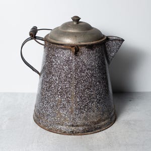 May include: An antique speckled enamelware coffee pot with a metal lid and handle. The pot has a dark brown and white speckled pattern, a spout, and a wooden handle grip. The pot is likely vintage and shows signs of wear.
