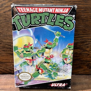 May include: A Teenage Mutant Ninja Turtles video game cartridge for the Nintendo Entertainment System. The box art features the four turtles in action poses, with a blue and purple background. The box is labeled "Teenage Mutant Ninja Turtles" and "Ultra Games".