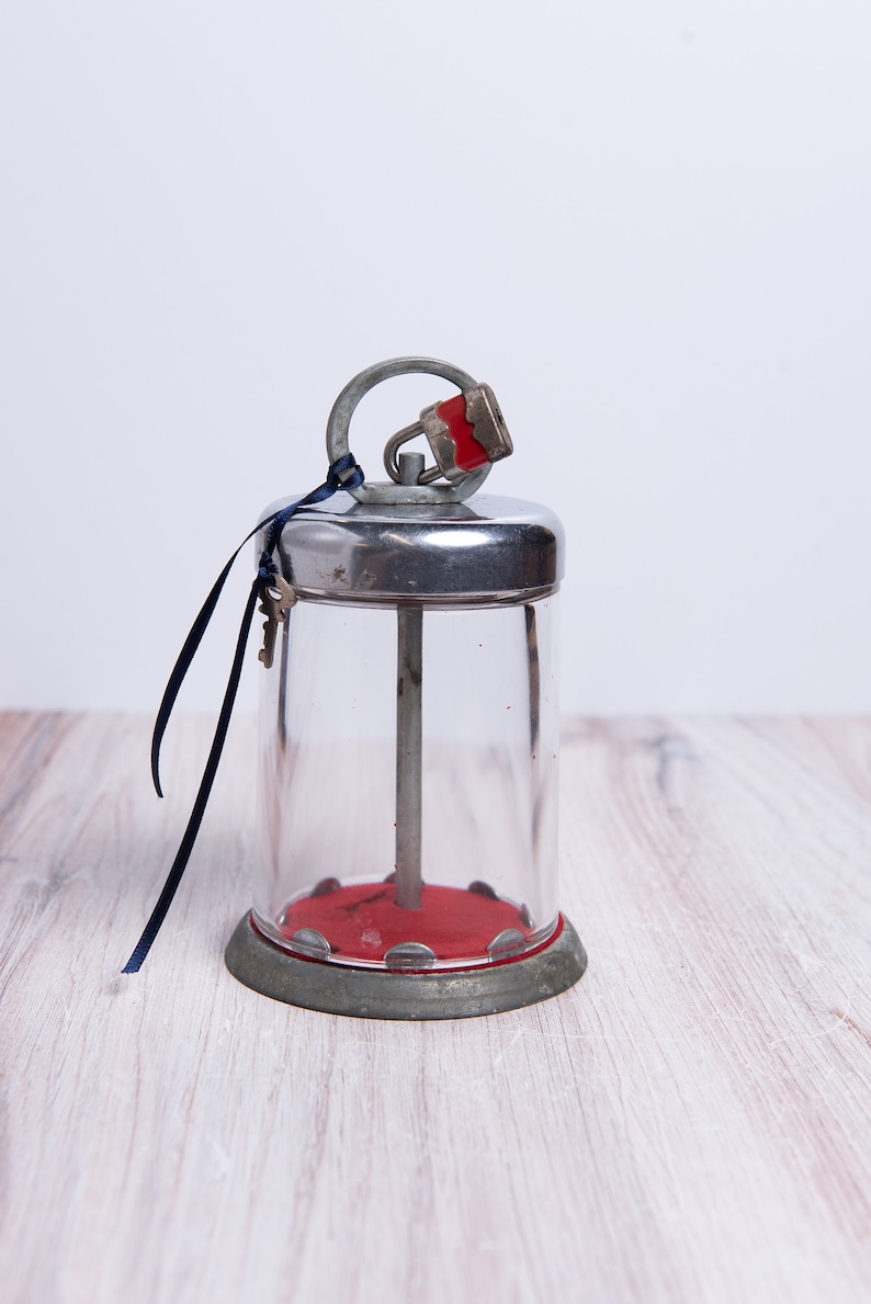 May include: A silver and glass jar with a red base and a padlock on the lid. The jar has a metal rod running through the center. A blue ribbon is tied around the lid.