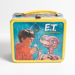 May include: Vintage E.T. The Extra-Terrestrial lunchbox. The lunchbox has a yellow frame and handle, with a blue background featuring E.T. and a boy. The lunchbox has the text "E.T. The Extra-Terrestrial".