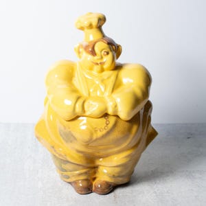 May include: A yellow ceramic cookie jar in the shape of a chef, with arms crossed. The chef wears a tall hat and apron. The jar has brown accents on the face and feet. The word "Food" is visible on the apron.