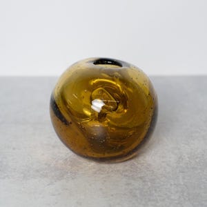 May include: A round, amber-colored glass vase with a small opening at the top. The vase has a bubble-like texture and a dark swirl design. The vase is a decorative item.