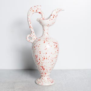 May include: A white ceramic pitcher with a unique handle and spout design. The pitcher is speckled with red paint, adding a decorative touch. The vase is approximately 12 inches tall.
