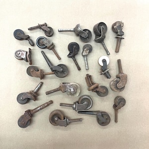 May include: A collection of antique metal casters, featuring various designs and sizes. The casters are primarily shades of brown and gray, showing signs of age and wear. Some have small wheels, while others have larger, more ornate designs. The collection is arranged on a neutral background.
