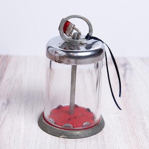May include: A vintage glass sugar dispenser with a metal lid and a red base. The dispenser has a metal rod that goes through the center and a small metal lock on the lid.