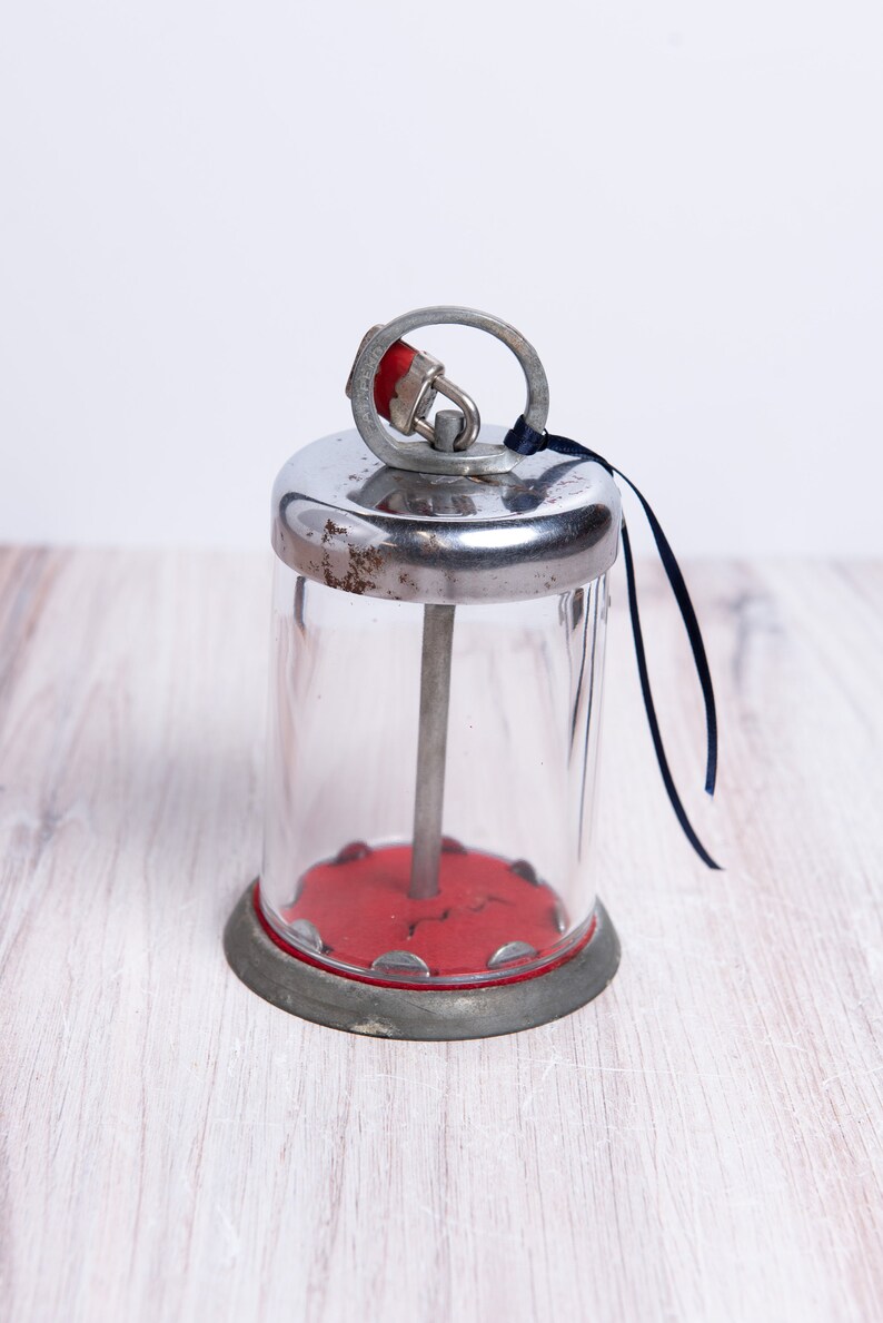 May include: A vintage glass sugar dispenser with a metal lid and a red base. The dispenser has a metal rod that goes through the center and a small metal lock on the lid.