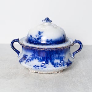 May include: A blue and white ceramic serving dish with a lid. The dish features a floral pattern and gold trim. It has two handles and a decorative finial. The dish is suitable for serving.