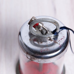 May include: A silver and red metal container with a loop and a blue string attached to the top. The container has a clear glass section and a red base.
