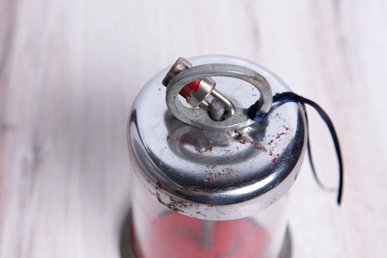 May include: A silver and red metal container with a loop and a blue string attached to the top. The container has a clear glass section and a red base.