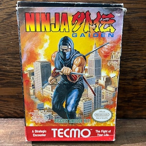 May include: A red, white, and black Nintendo game cartridge box for Ninja Gaiden. The box features a ninja character in a blue suit holding a sword, standing in front of a city skyline. The text on the box reads "Ninja Gaiden" and "Biggest Arcade Hit!"