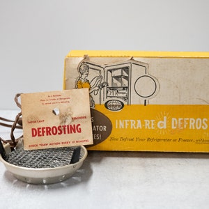 May include: A yellow box with the words "Infra-Red Defroster" and a picture of a refrigerator. The box is next to a small, silver, metal defroster with a black handle and a cord.